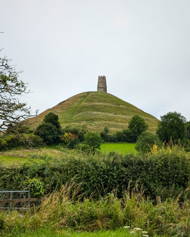 View of Glastonbury Tor from a distance with the church tower on top of the hilll, and hedges and fields in the foreground