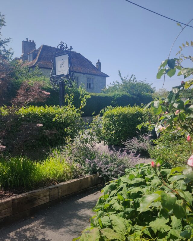 Photo of a country inn sign taken from the inn's garden. Bushes, shrubs and flowers can be seen in the foreground and a traditional stone cottage is in the background