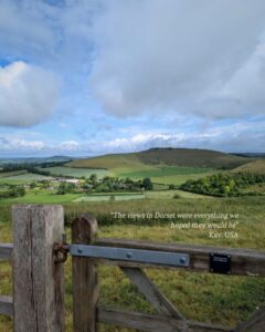 View out over the lush green countryside of Dorset. A wooden gate is in the foreground and rolling hills in the background. Next to the gate is a testimonial: “The views in Dorset were everything we hoped they would be” Kay, USA