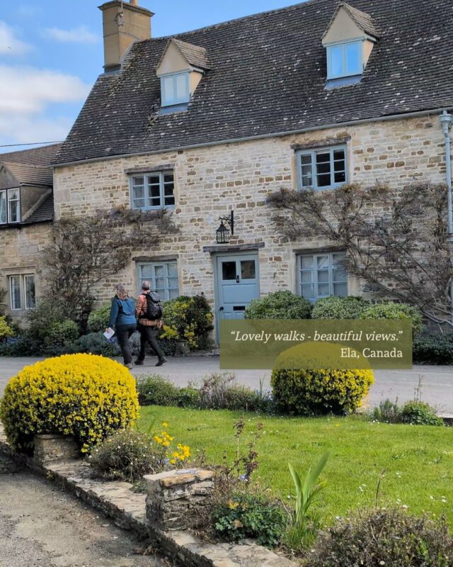 A couple walk past a row of old stone Cotswolds cottages behind a village green. A guest testimonial is positioned in the middle right of the photo: “Lovely walks - beautiful views.” Ela, Canada