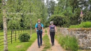 Two hikers walk down a gravel path in the countryside. The words 'A day in the life of a Foot Trail are shown in the top left corner.