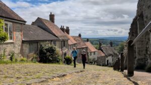 Two hikers are walking up a steep cobbled hill with quaint thatched and tiled roof cottages descending down the hill