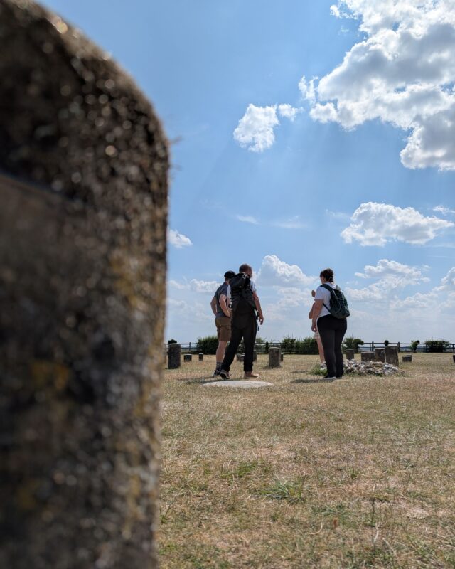 A group of people stand around an information board in a field. There are lots of small concrete posts in the ground marking where the pillars of Woodhenge would have stood.