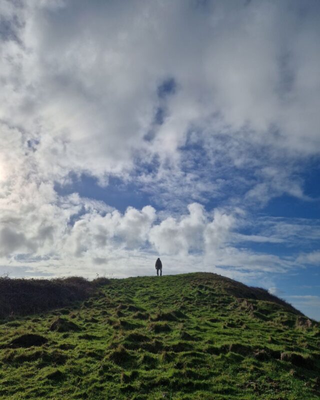 A long figure stands on a hill in the distance silhouetted again a blue sky with white clouds.
