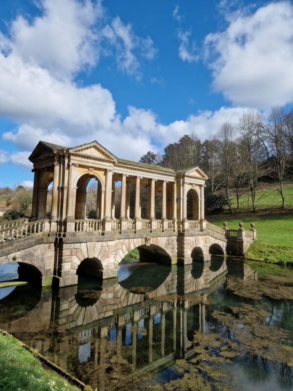 View of Prior Park Palladian Bridge in Bath on a bright spring day. The ornate bridge is mirrored in the still water below.