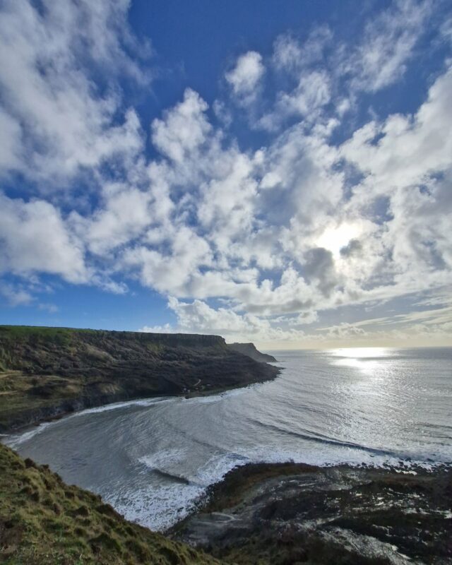 View from a cliff top over a secluded bay, with the coastline stretching off into the distance and the rippled sea stretching out to the right.