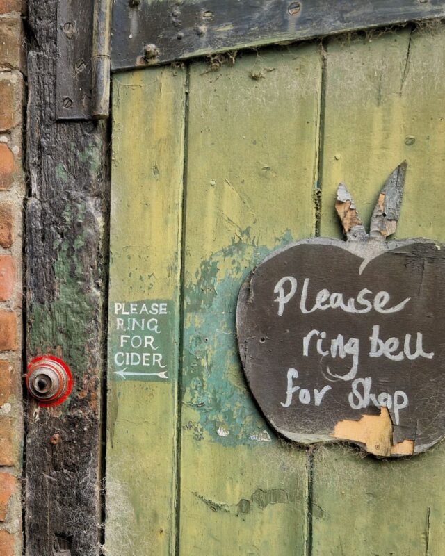 An old wooden door with paint peeling off it. To the left is a doorbell with the sign 'Ring for Cider'