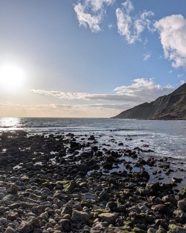 View over a calm sea from a pebble beach. There is a cliff to the right, and on the left, the evening sun is just above the horizon.