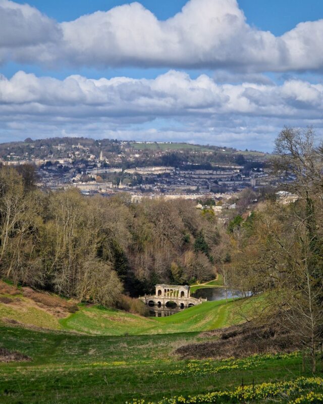 View over landscaped gardens with a palladian bridge over a river in middleground and the city of Bath stretching out behind park trees