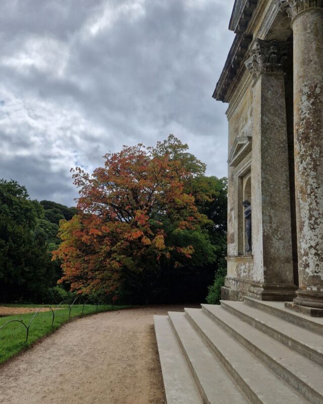 A path winds its way through an English country house estate past the entrance to a Roman-style temple with steps leading up to stone columns