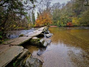 View of an old stone clapper bridge extending out over a river in an autumnal landscape