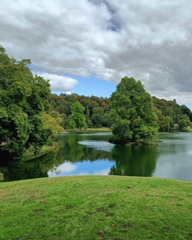 View of a lake in landscaped country house gardens with lush green trees in the background and a tree in the centre of the lake.