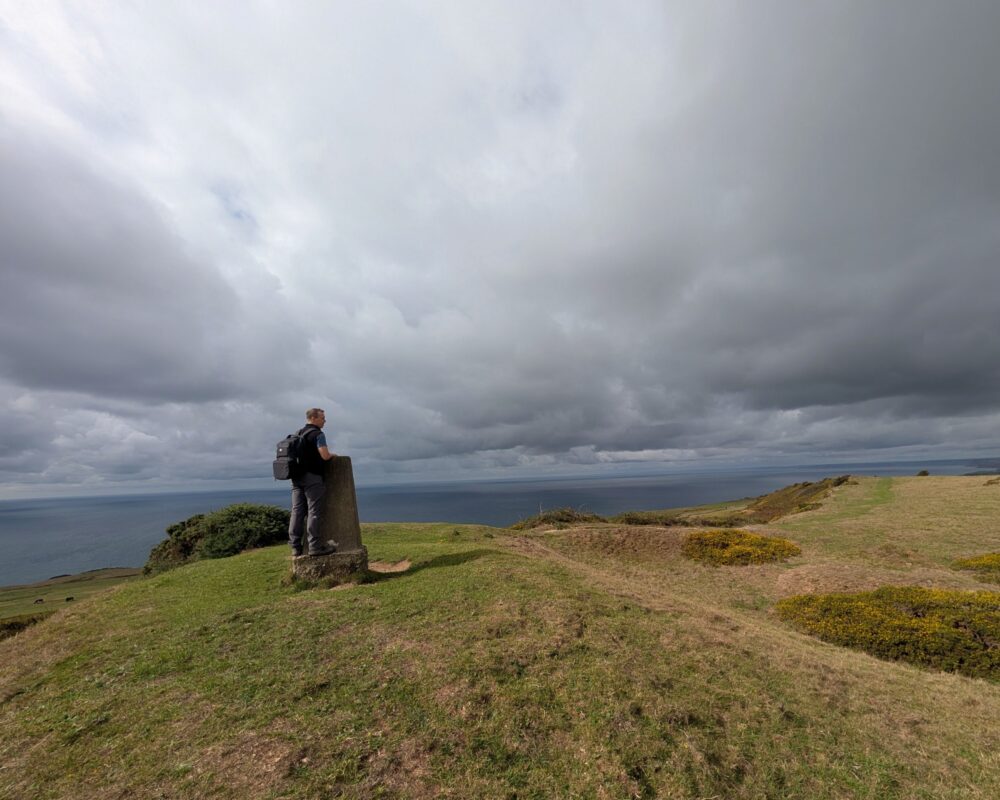 A lone hiker stands on top of a hill next to a trig point. The undulating landscape stretches off to the right while the sea stretches off to the left under an overcast sky.