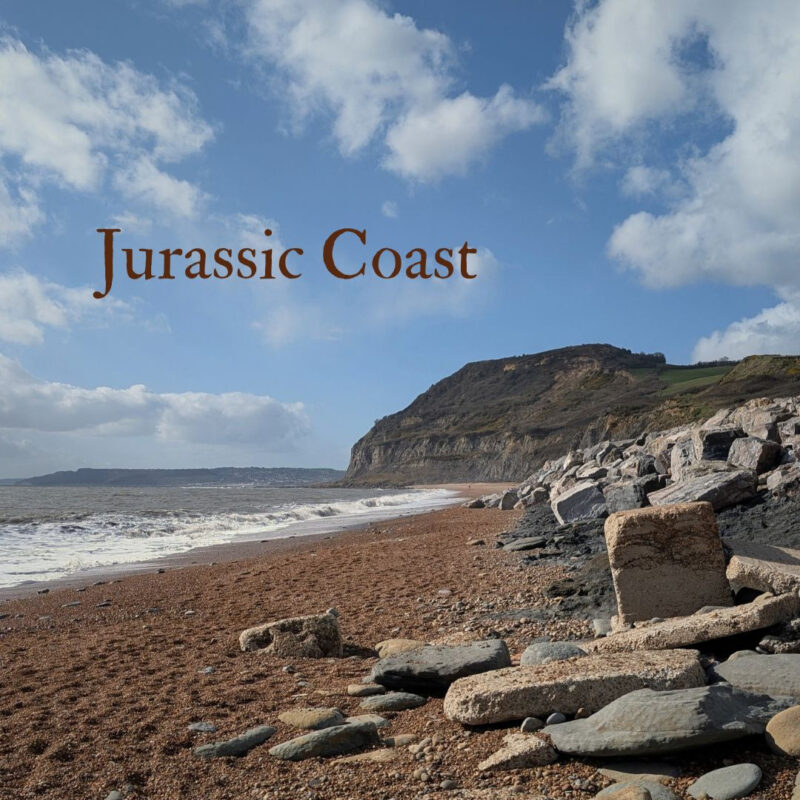 A view of the Dorset coast with rocks and a pebble beach in the foreground and Golden Cap in the background. The words Jurassic Coast are shown in the top left corner.