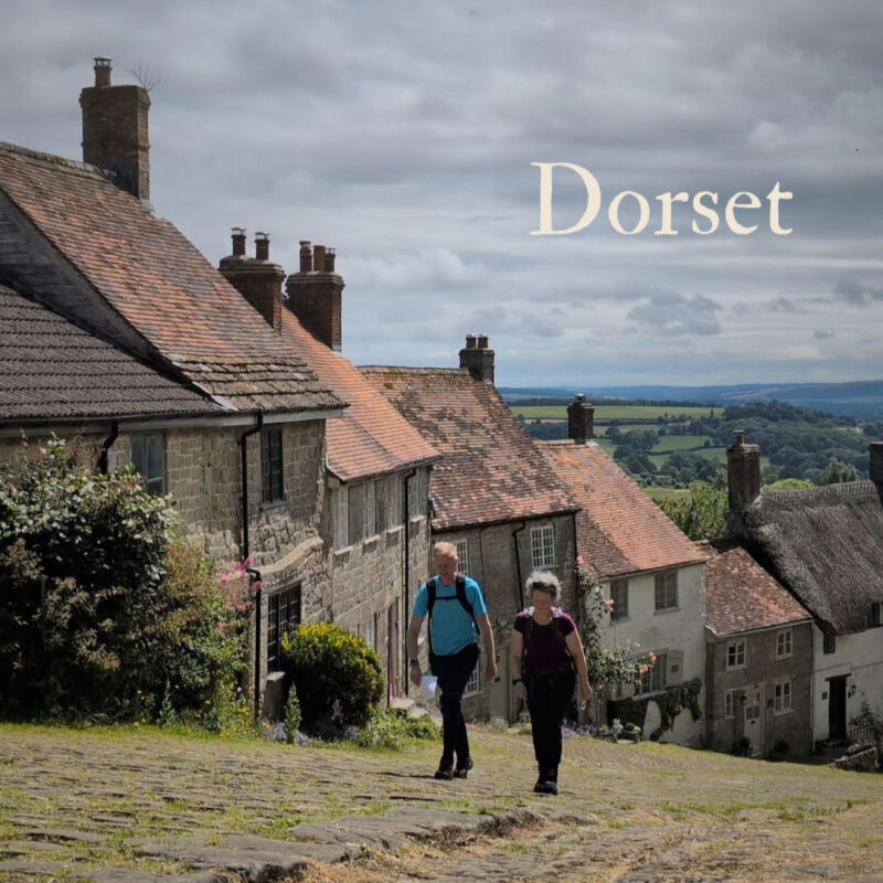 Two people walking up a steep cobbled street lined with picturesque cottages. The word Dorset is shown in the top right corner.