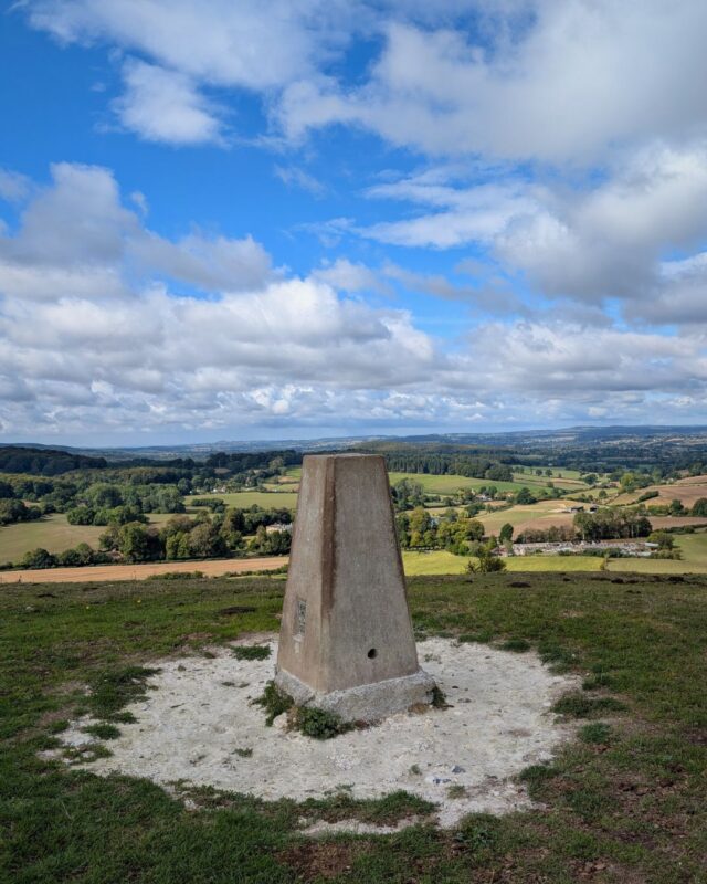 View of a trig point on top of a hill with the countryside stretching out into the distance beyond under a bright blue sky and white fluffy clouds.