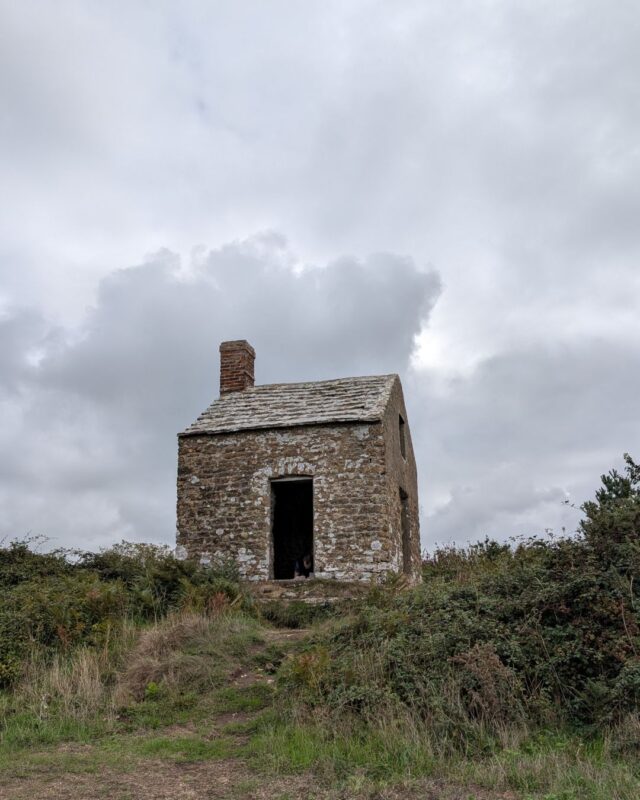 An small, grey abandoned stone cottage on top of a hill silhouetted against a grey sky. It has a grey slate roof and chimney and a doorway with no door.