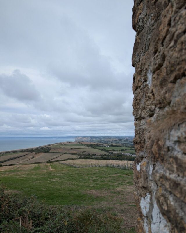 A view over undulating coastal landscape from a hill. To the right of the image is an old stone wall while the countryside and sea can be seen stretching out into the distance.