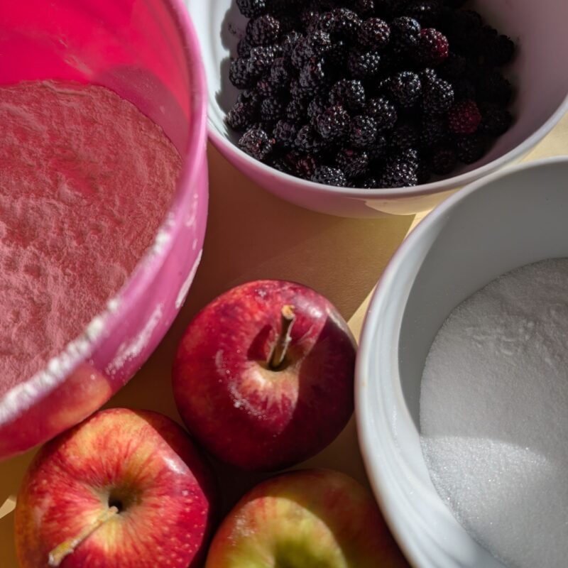 Three mixing bowls filled with flour, sugar and blackberries and three apples in between the bowls - some of the ingredients for apple &blackberry crumble
