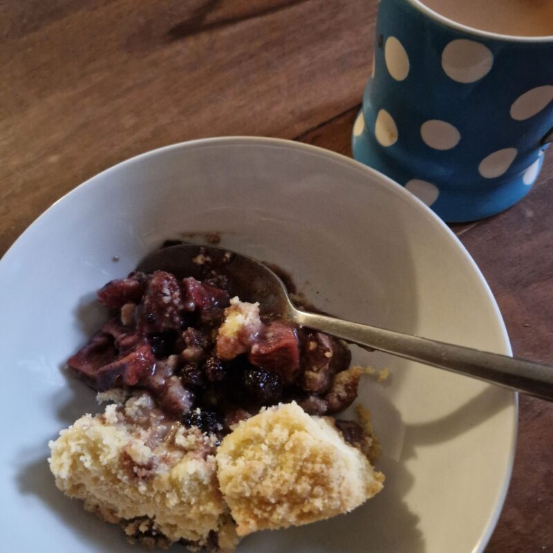 A portion of apple & blackberry crumble in a bowl with a spoon. A cup of tea in a blue and white mug is next to the bowl.
