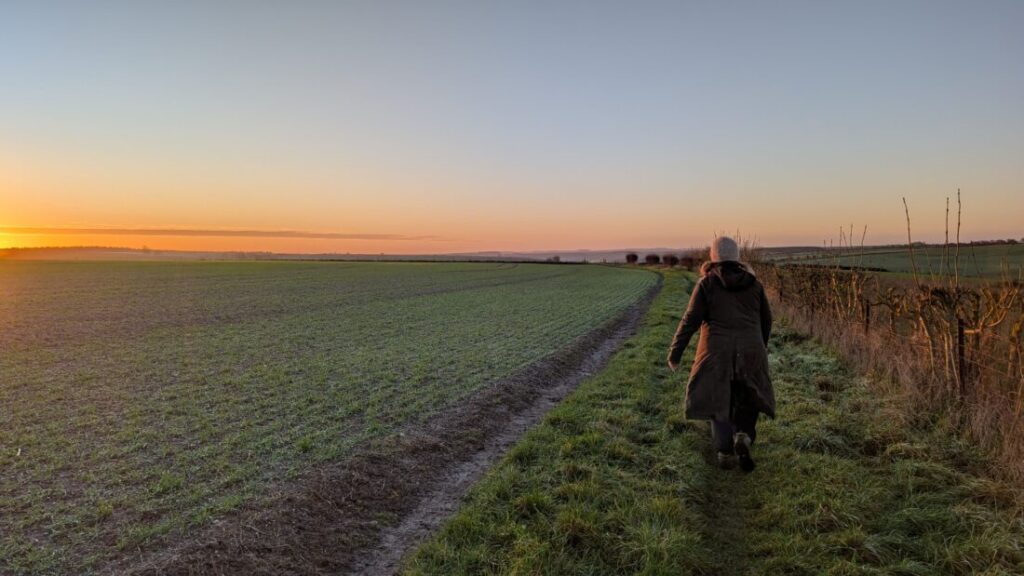 Foot Trails Co-Founder Alison walks along the edge of a frosty field at dawn with the first signs of the sunrise showing to the left.