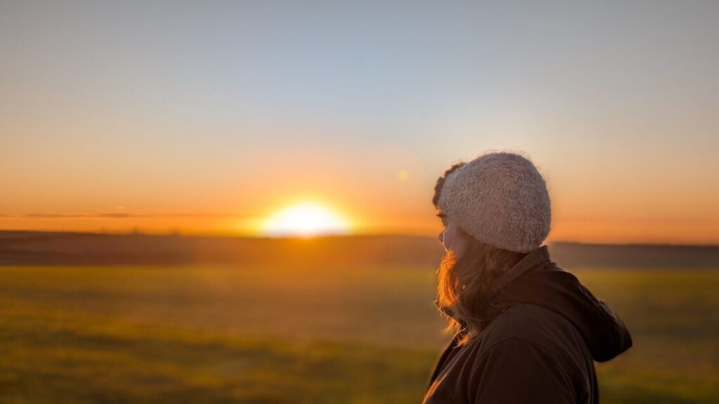 View of Foot Trails Co-Founder Alison's head and shoulders from the side. Alison is looking at the sun rising over a field.