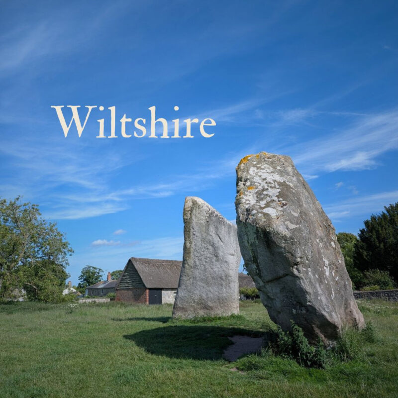 Two large standings stones in a field with a cottage just visible in the background. The word Wiltshire is displayed in the top left corner.