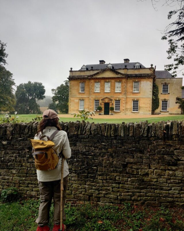 A hiker stands in fonrt of a hedge with their back to the camera looking at a grand country house
