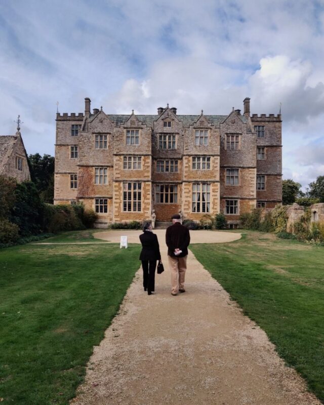 Two people are walking down an grand driveway towards a beautiful Jacobean house