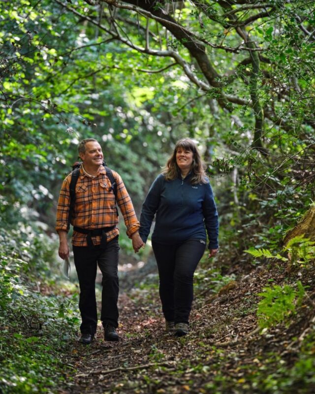 Foot Trails' Founders Alison and David are walking through a woodland holding hands and smiling at the camera.