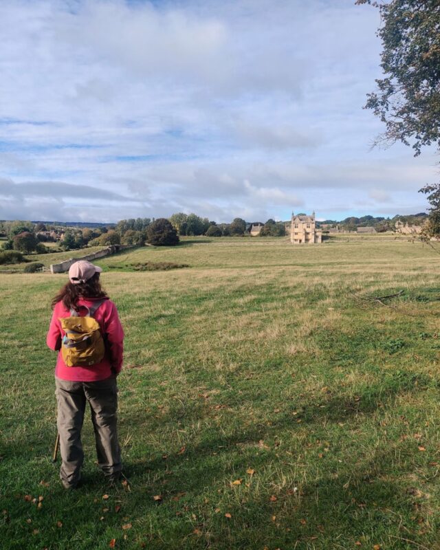 A female hiker is standing in a field with her back to the camera. She is looking out ta a country house in the distance.