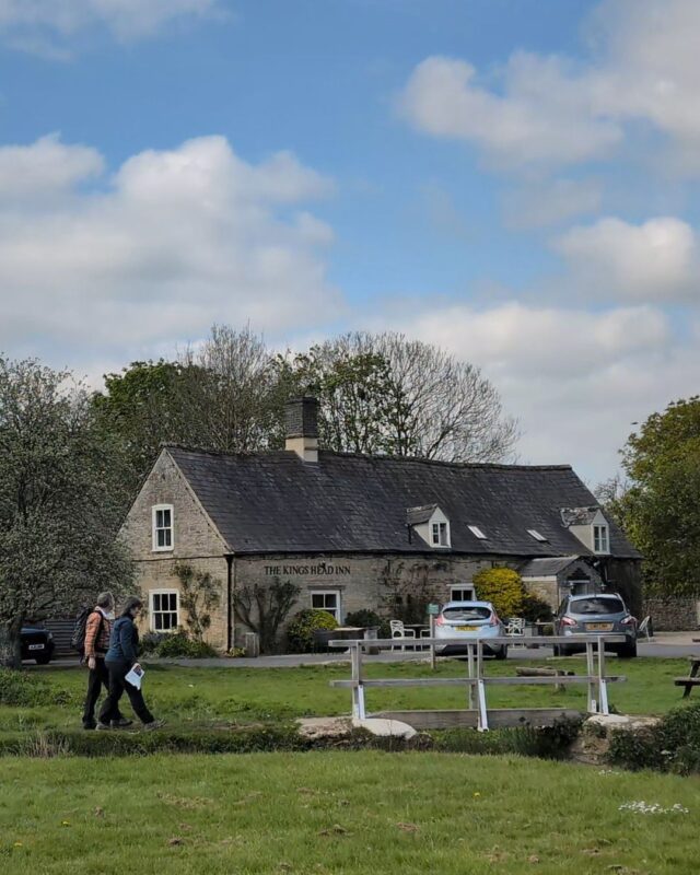 View of a Cotswolds country inn with two hikers walking across a footbridge on the green in front of the pub