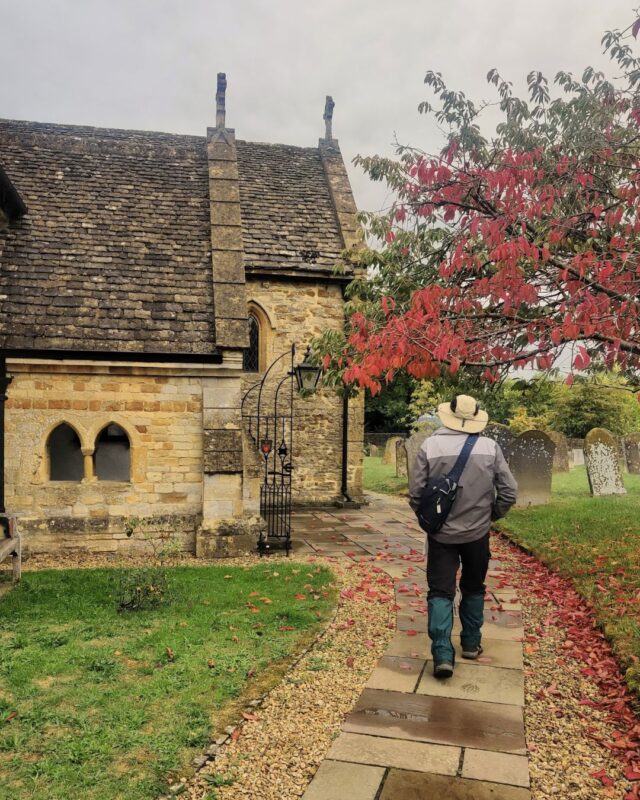 A hiker walks through a church graveyard towards an ancient church under a tree with bright red leaves