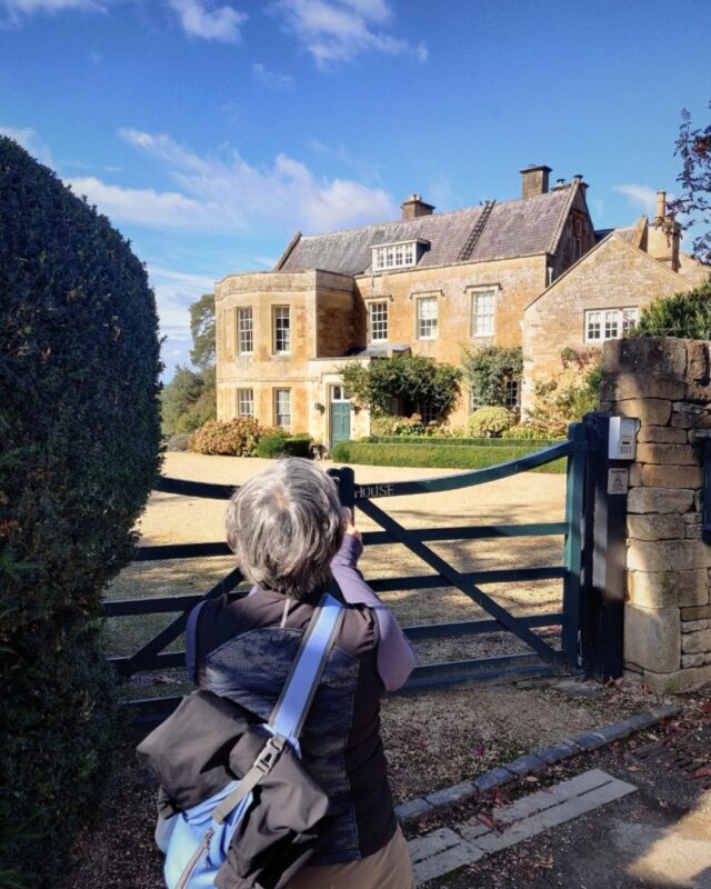 A female hiker is taking a photo of a grand Cotswolds country house.