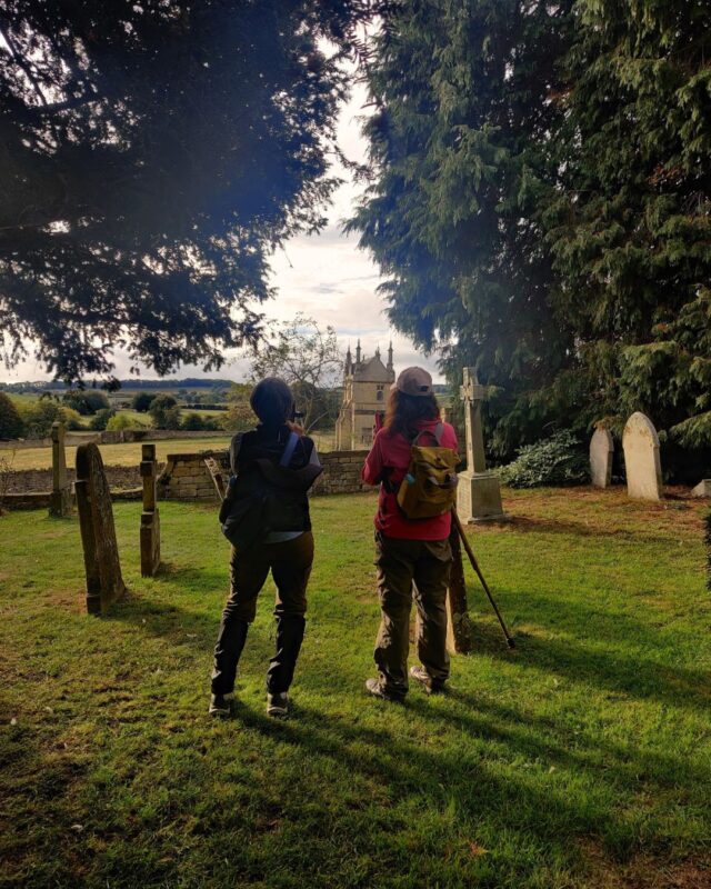 Two hikers stand in a graveyard under some trees. They have their backs to the camera and are looking at a grand country house on the other side of the cemetery wall.
