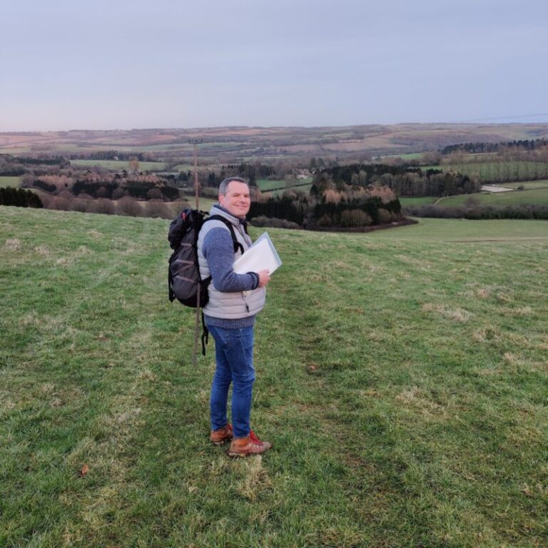 Foot Trails Founder David Howell is standing in a field in winter, warmly dressed in hiking gear and holding some trail notes.
