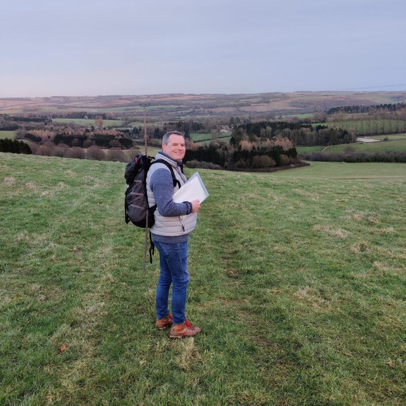 Foot Trails Founder David Howell is standing in a field in winter, warmly dressed in hiking gear and holding some trail notes.