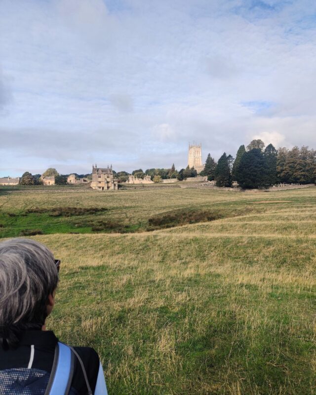 A female hiker stands with her back to the camera looking out over rolling fields to the banqueting hall at Chipping Campden.